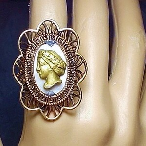 May include: A gold-toned ring with an oval cameo featuring a profile of a woman with a laurel wreath. The cameo is set against a white background, framed by a decorative, openwork border. The ring is on a mannequin hand with red nail polish.