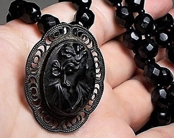 May include: A black glass cameo pendant necklace with a detailed design of a woman's face, set in a silver-toned filigree frame. The necklace is made of black faceted beads.
