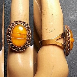 May include: A gold ring with an oval-shaped amber stone set in a decorative frame. The ring is worn on a finger with red nail polish.