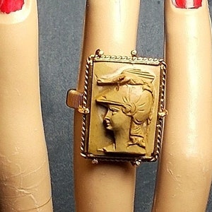 May include: A gold ring with a cameo featuring a woman's profile wearing a helmet. The cameo is set in a square frame with a rope-like border.