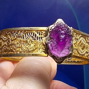 Könnte beinhalten: Ein goldenes Armband mit einem verzierten Design und einem violetten Kamee mit einem Frauenkopf.