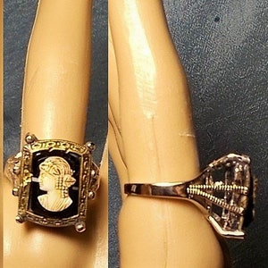 Op de afbeelding: Een gouden ring met een zwarte cameo met het profiel van een vrouw. De cameo is gezet in een gouden frame met een zwarte achtergrond. De ring wordt aan een vinger gedragen.