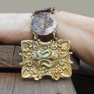 May include: A gold-toned bracelet with a large, ornate pendant featuring a green, red, and gold design. The pendant is attached to a chain with a smaller, round pendant featuring a carved design.
