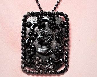 May include: A black necklace with a large, square pendant featuring a cameo of a woman's face surrounded by black beads.
