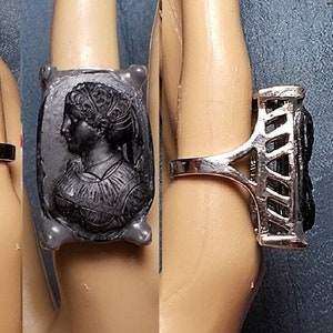 May include: A silver ring with a black cameo featuring a woman's profile. The cameo is set in a rectangular frame with a decorative border.