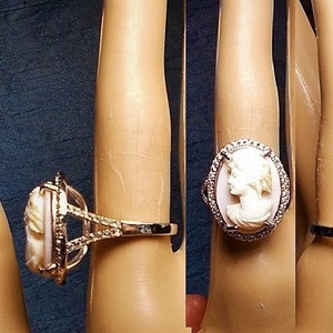 May include: A cameo ring featuring a white profile set against a pink background, surrounded by small clear stones. The ring has a gold-colored band and a decorative setting. The ring is displayed on a mannequin finger.