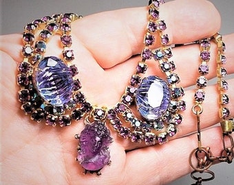 May include: A vintage purple and iridescent rhinestone necklace with a cameo pendant. The necklace features two large, faceted stones and a smaller cameo pendant with a carved design. The necklace is made of gold-tone metal and has a lobster claw clasp.