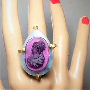 Puede incluir: Un anillo con un camafeo morado de perfil de mujer engastado en una piedra blanca con detalles dorados.