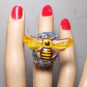 May include: A yellow and black bee ring with blue and white details. The ring is on a mannequin hand.