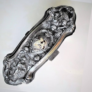May include: A silver-plated trinket box with an ornate design featuring cherubs and floral motifs. The box has a heart-shaped center with a raised image of a couple.
