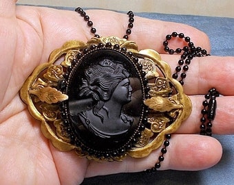 May include: A gold-tone pendant necklace with a black cameo featuring a woman's profile. The cameo is surrounded by a decorative frame with black beads. The necklace is on a black chain.
