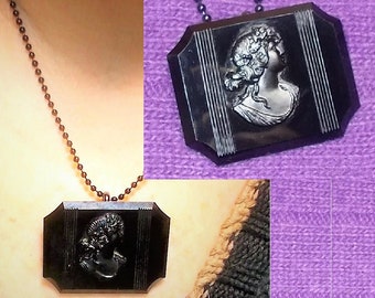 May include: A black cameo necklace with a detailed portrait of a woman with a floral headpiece. The cameo is set in a black octagonal frame and hangs from a black chain.