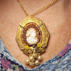 Può includere: Una collana d'oro con un ciondolo a cammeo che presenta il profilo di una donna. Il ciondolo è circondato da perline marroni e gialle e ha un grappolo di perle bianche che pendono dal fondo.