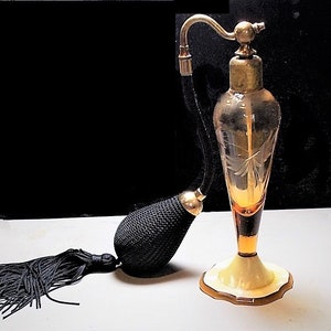 May include: An amber glass perfume bottle with a clear glass top and a black tassel. The bottle has an etched design and sits on a white base.