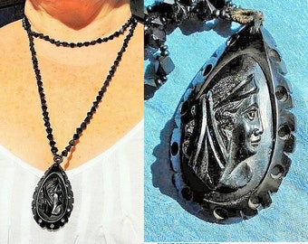 May include: A black beaded necklace with a carved black pendant featuring a woman's face. The pendant is shaped like a teardrop and has a detailed design.