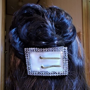 May include: A white rectangular hair clip with two gold metal bars and a border of clear rhinestones.