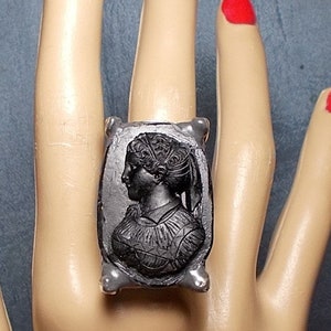 May include: A silver ring with a black cameo featuring a woman's profile. The cameo is set in a rectangular frame with a textured surface.