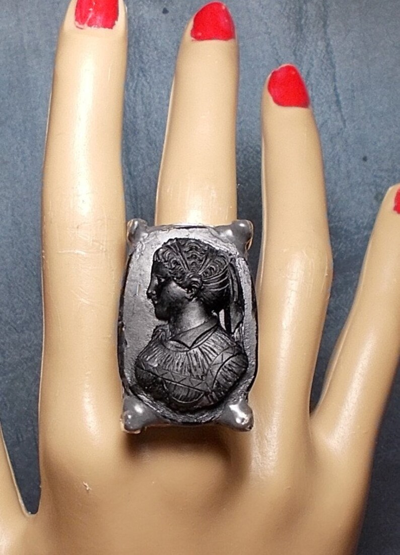 May include: A silver ring with a black cameo featuring a woman's profile. The cameo is set in a rectangular frame with a textured surface.