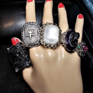 May include: Five silver rings with different designs. The rings feature a compass, a cameo, a black cameo, a black cameo with a purple stone, and a pink stone with green stones.