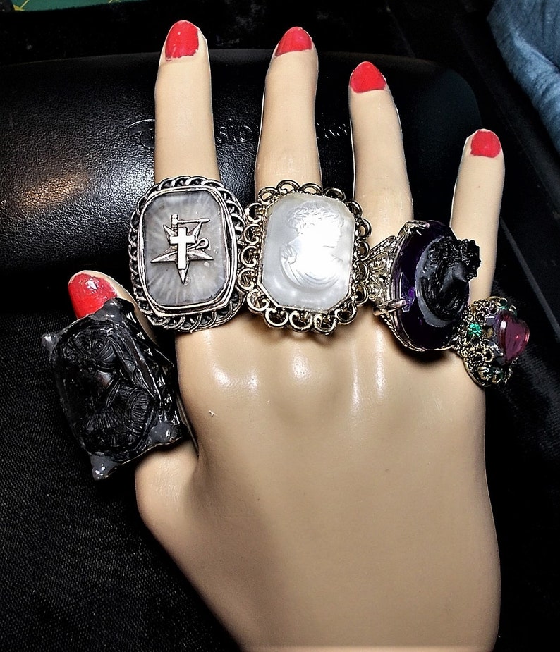 May include: Five silver rings with different designs. The rings feature a compass, a cameo, a black cameo, a black cameo with a purple stone, and a pink stone with green stones.