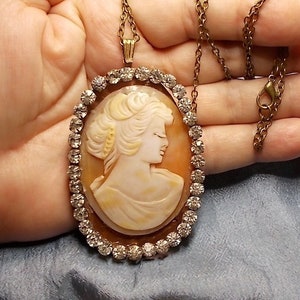 May include: Oval cameo pendant necklace featuring a profile of a woman with a cream-colored face and hair. The cameo is set in a gold-toned frame with a border of clear rhinestones. The necklace has a gold-toned chain.