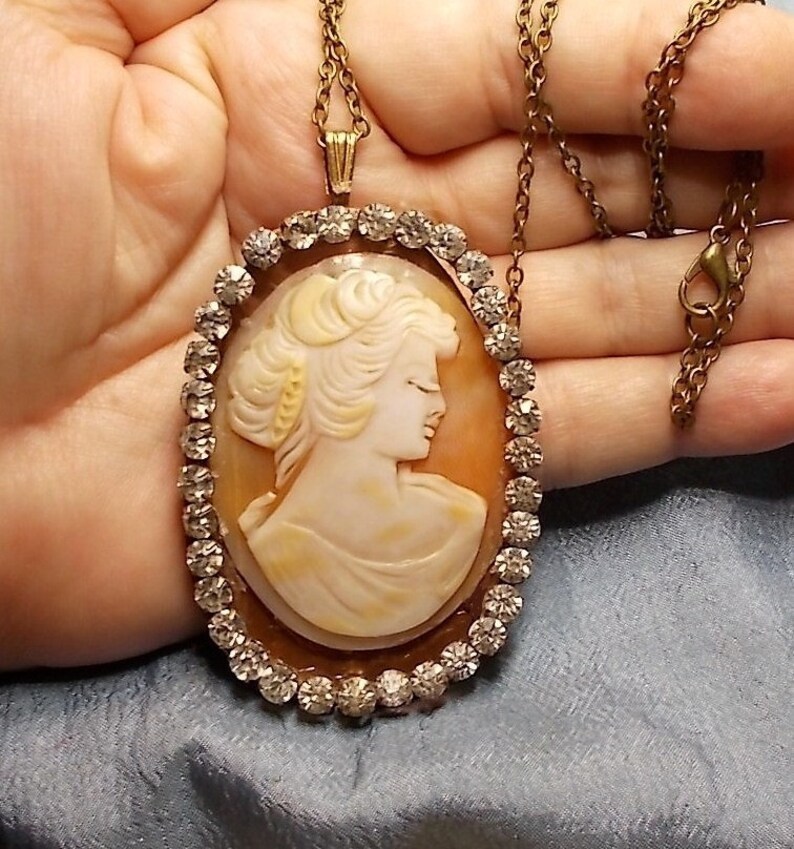 May include: Oval cameo pendant necklace featuring a profile of a woman with a cream-colored face and hair. The cameo is set in a gold-toned frame with a border of clear rhinestones. The necklace has a gold-toned chain.