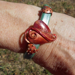 May include: A green leather bracelet with a copper heart-shaped charm and gold accents. The charm has a swirl design and is attached to the bracelet with a metal loop.