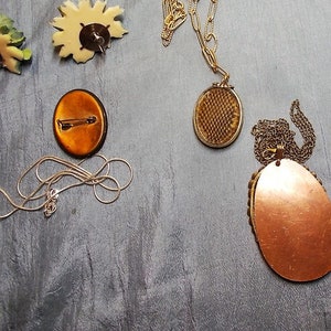 May include: An assortment of vintage jewelry pieces, including a gold-toned locket necklace, a silver chain necklace, and a copper-colored oval pendant necklace. Also, a floral brooch and a pin with a copper-colored oval design.
