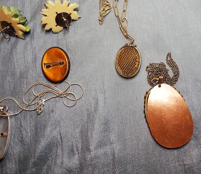 May include: An assortment of vintage jewelry pieces, including a gold-toned locket necklace, a silver chain necklace, and a copper-colored oval pendant necklace. Also, a floral brooch and a pin with a copper-colored oval design.