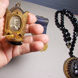 May include: A gold locket with a black cameo and a photo of a man inside. The locket is open and the inside is lined with black velvet. The locket is hanging from a black beaded necklace.