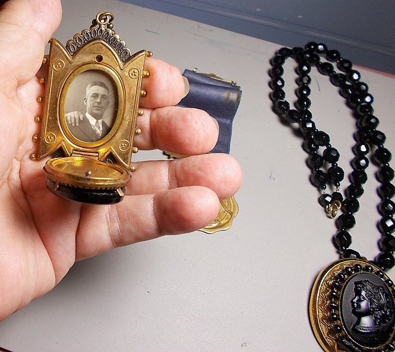 May include: A gold locket with a black cameo and a photo of a man inside. The locket is open and the inside is lined with black velvet. The locket is hanging from a black beaded necklace.