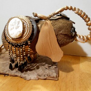 May include: A gold and brown elephant figurine with a cameo and beaded trim. The elephant has a long trunk and large ears. The figurine is sitting on a wooden surface.
