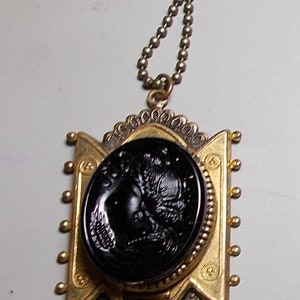 May include: A gold pendant necklace with a black cameo featuring a detailed profile of a woman's face. The pendant is surrounded by a decorative gold frame and hangs from a gold chain.
