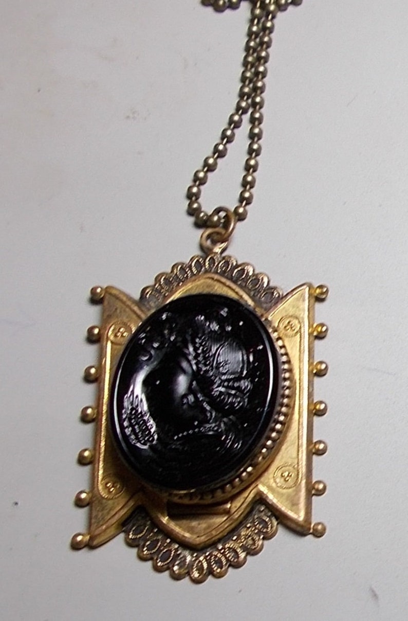 May include: A gold pendant necklace with a black cameo featuring a detailed profile of a woman's face. The pendant is surrounded by a decorative gold frame and hangs from a gold chain.