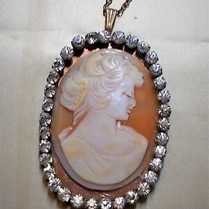 May include: Oval pendant necklace featuring a cameo of a woman in profile. The cameo is set in a copper-colored frame and surrounded by a border of clear, sparkling stones. The necklace has a delicate gold-colored chain.