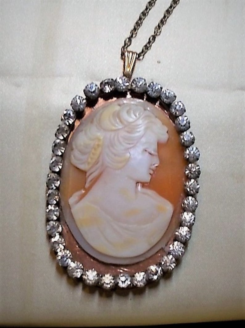 May include: Oval pendant necklace featuring a cameo of a woman in profile. The cameo is set in a copper-colored frame and surrounded by a border of clear, sparkling stones. The necklace has a delicate gold-colored chain.