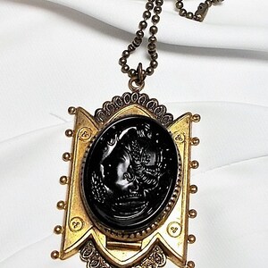 May include: A gold tone necklace with a black cameo pendant featuring a woman's profile. The pendant is surrounded by a decorative frame and the chain is made of small round beads.