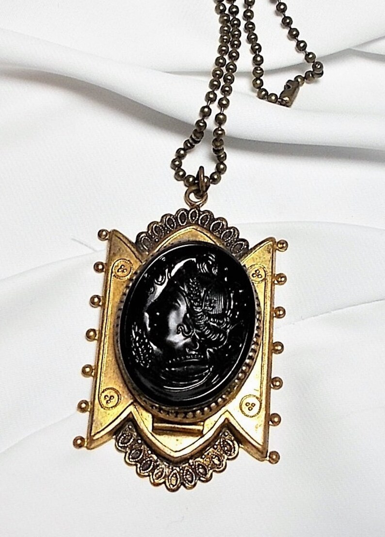 May include: A gold tone necklace with a black cameo pendant featuring a woman's profile. The pendant is surrounded by a decorative frame and the chain is made of small round beads.