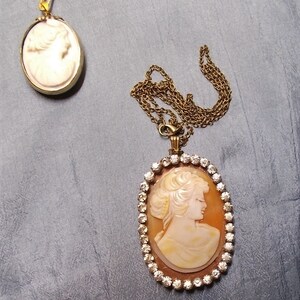 May include: Two cameo necklaces. One is a small, oval pendant with a gold-colored frame and chain. The other is a larger, oval pendant with a woman's profile, surrounded by clear crystals, on a gold-colored chain.