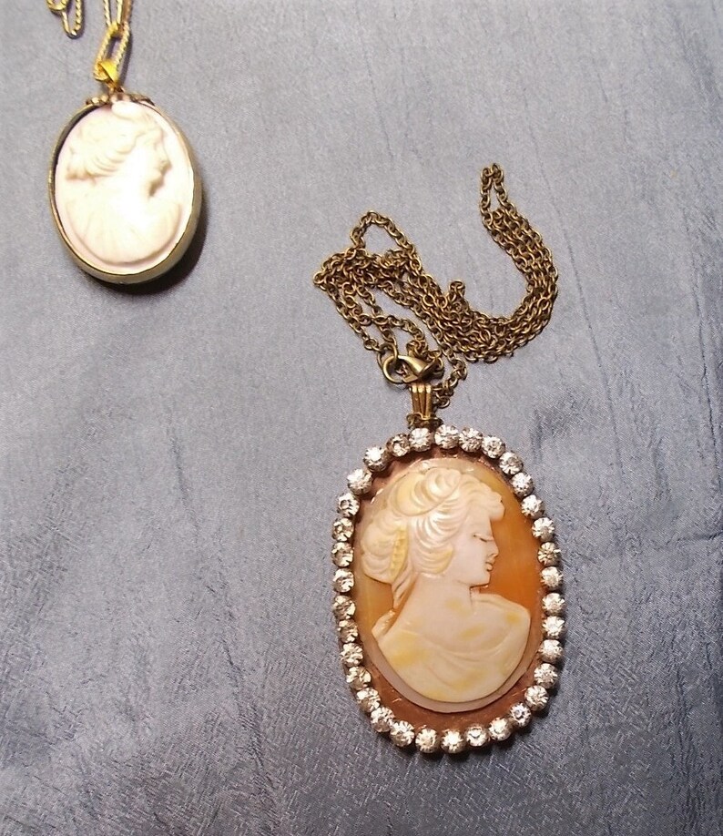 May include: Two cameo necklaces. One is a small, oval pendant with a gold-colored frame and chain. The other is a larger, oval pendant with a woman's profile, surrounded by clear crystals, on a gold-colored chain.