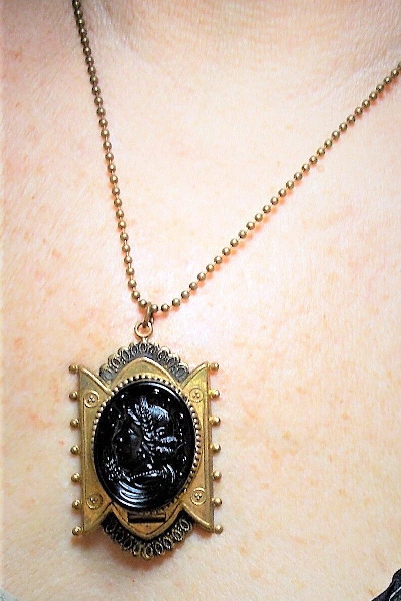 May include: A gold chain necklace with a black cameo pendant. The pendant is set in a gold frame with intricate details. The cameo features a woman's profile.
