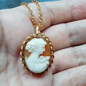 Puede incluir: Un collar de cadena dorada con un colgante de camafeo que presenta el perfil de una mujer en blanco sobre un fondo marrón.