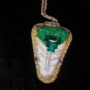 Könnte beinhalten: Eine goldene Kettenkette mit einem Anhänger, der einen großen Smaragd-Edelstein zeigt, der von kleineren Smaragden umgeben ist und in einen goldenen Rahmen gefasst ist.