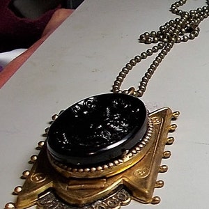 May include: A gold pendant necklace with a black cameo featuring a detailed design. The pendant is attached to a gold chain with small round beads.