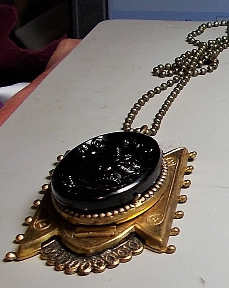 May include: A gold pendant necklace with a black cameo featuring a detailed design. The pendant is attached to a gold chain with small round beads.