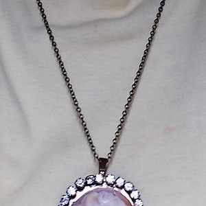 May include: A cameo pendant necklace featuring a white profile portrait set against an orange background. The oval pendant is framed with clear crystals and hangs from a dark chain.