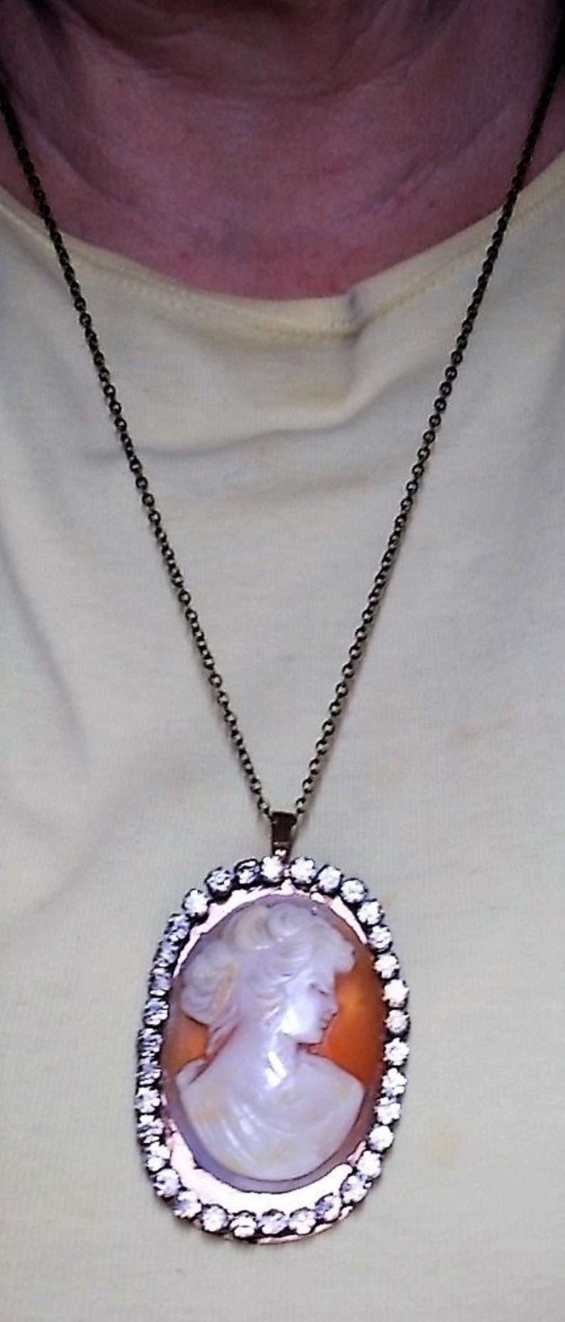 May include: A cameo pendant necklace featuring a white profile portrait set against an orange background. The oval pendant is framed with clear crystals and hangs from a dark chain.