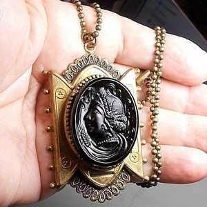 May include: A gold tone pendant necklace with a black cameo featuring a woman's profile. The pendant is ornate and has a detailed design. The chain is a simple ball chain.