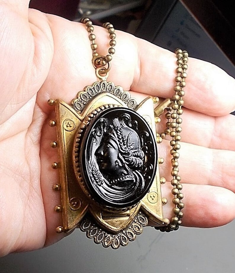 May include: A gold tone pendant necklace with a black cameo featuring a woman's profile. The pendant is ornate and has a detailed design. The chain is a simple ball chain.