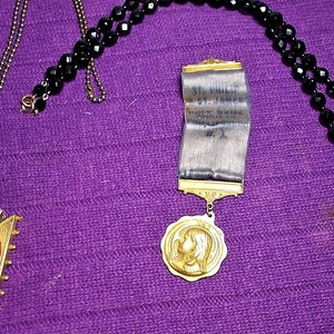 May include: Three necklaces with black beads and gold pendants. One pendant features a black cameo of a woman's profile, another has a gold medal with a woman's profile, and the third has a black cameo of a woman's profile in a gold frame with a ribbon that reads "St. Hilda's Guild, 1900, New York, N.Y."
